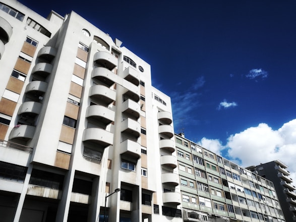A view of a residential building with a modern architectural design featuring rounded balconies. The sky is bright blue with some white clouds, creating a vibrant contrast. The building is mostly white with sections of tan-colored wall panels.
