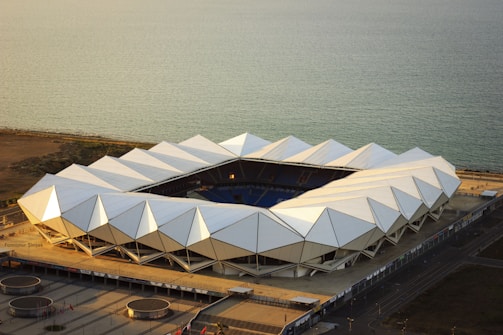 A geometric stadium with a unique, angular white roof structure is situated near the coastline. The surrounding area appears open, leading to the blue expanse of the sea, which stretches out to the horizon. The stadium is illuminated by warm sunlight, creating a striking contrast with the dark seating inside.
