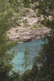 A scenic view featuring clear turquoise waters surrounded by lush green trees and rocky cliffs. In the background, several rustic buildings are nestled among the rugged hillside, providing a sense of seclusion and tranquility.