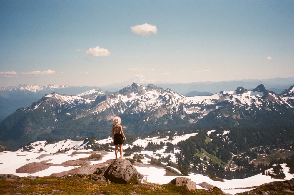 woman in white shirt and black pants standing on rock near snow covered mountains during daytime