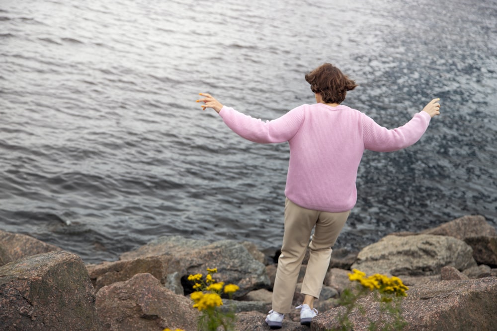 girl in pink long sleeve shirt and brown pants standing on rock near body of water