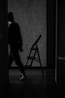 A blurred silhouette of a person walking past a stepladder in a dimly lit room, creating a sense of motion and mystery.