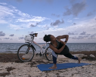A person is performing a yoga pose on a blue mat by the seaside with a bicycle parked nearby. The sky is partly cloudy, and the sea is calm.