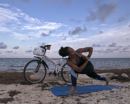 A person is performing a yoga pose on a blue mat by the seaside with a bicycle parked nearby. The sky is partly cloudy, and the sea is calm.