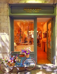 A quaint bakery storefront with a sign reading 'Martina Sweet Cakes' features a charming blue bicycle adorned with baskets of vibrant flowers parked outside. Inside, the warm glow highlights the decorative interior and pastries on display.