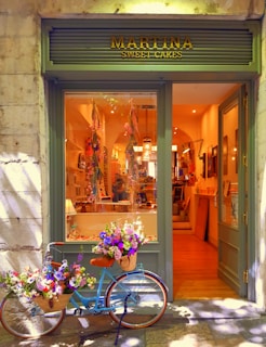 A quaint bakery storefront with a sign reading 'Martina Sweet Cakes' features a charming blue bicycle adorned with baskets of vibrant flowers parked outside. Inside, the warm glow highlights the decorative interior and pastries on display.
