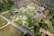 Aerial view of a rural residential area with several houses, roads, and patches of forested land. The landscape includes well-maintained lawns, driveways, and a small pond near one of the properties. Trees and grassy areas dominate the scene, creating a blend of natural and developed land.