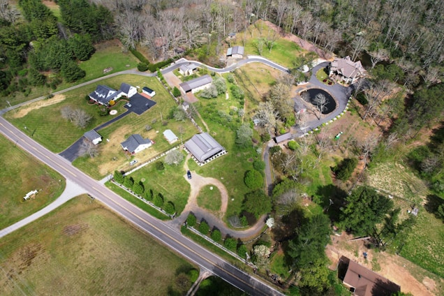 Aerial view of a rural residential area with several houses, roads, and patches of forested land. The landscape includes well-maintained lawns, driveways, and a small pond near one of the properties. Trees and grassy areas dominate the scene, creating a blend of natural and developed land.