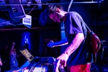 A musician is immersed in a live performance, adjusting equipment on a table with a laptop and mixer, creating an energetic atmosphere with vibrant blue lighting. People in the background add to the lively setting, suggesting a club or concert environment.