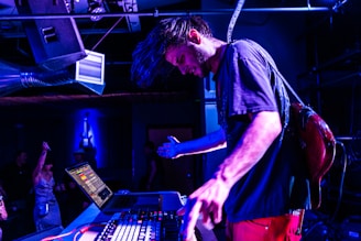 A musician is immersed in a live performance, adjusting equipment on a table with a laptop and mixer, creating an energetic atmosphere with vibrant blue lighting. People in the background add to the lively setting, suggesting a club or concert environment.