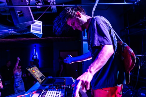 A musician is immersed in a live performance, adjusting equipment on a table with a laptop and mixer, creating an energetic atmosphere with vibrant blue lighting. People in the background add to the lively setting, suggesting a club or concert environment.