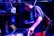 A musician is immersed in a live performance, adjusting equipment on a table with a laptop and mixer, creating an energetic atmosphere with vibrant blue lighting. People in the background add to the lively setting, suggesting a club or concert environment.