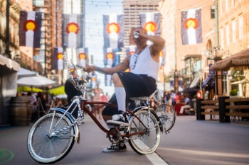 A person casually rides a lowrider bicycle down a busy urban street lined with brick buildings and adorned with multiple flags featuring a distinctive circular emblem. The rider is dressed in a white tank top and shorts, and is drinking from a can. Overhead string lights add to the festive, leisurely atmosphere.