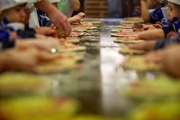 A group of children and adults are gathered around a long table, each preparing and assembling pizzas. The focus is on the many small hands working with dough and toppings. The scene suggests a fun and collaborative cooking activity.