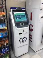 A white cryptocurrency ATM machine with a touchscreen display stands next to a refrigerated display in a convenience store. The screen shows options for buying Bitcoin with different amounts. Beside the ATM, shelves are stocked with snack items including Cheetos and Doritos.