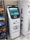 A white cryptocurrency ATM machine with a touchscreen display stands next to a refrigerated display in a convenience store. The screen shows options for buying Bitcoin with different amounts. Beside the ATM, shelves are stocked with snack items including Cheetos and Doritos.