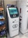 A white cryptocurrency ATM machine with a touchscreen display stands next to a refrigerated display in a convenience store. The screen shows options for buying Bitcoin with different amounts. Beside the ATM, shelves are stocked with snack items including Cheetos and Doritos.