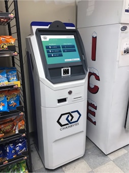A white cryptocurrency ATM machine with a touchscreen display stands next to a refrigerated display in a convenience store. The screen shows options for buying Bitcoin with different amounts. Beside the ATM, shelves are stocked with snack items including Cheetos and Doritos.