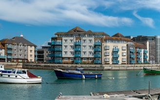 A waterfront scene featuring several modern residential buildings with blue balconies located alongside a marina. In the foreground, a few boats are anchored in calm water, with a clear blue sky featuring wispy clouds in the background.