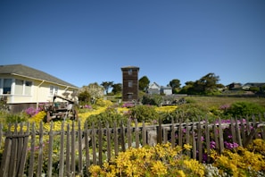 A charming homestead with a rustic wooden fence and blooming garden under a bright blue sky.