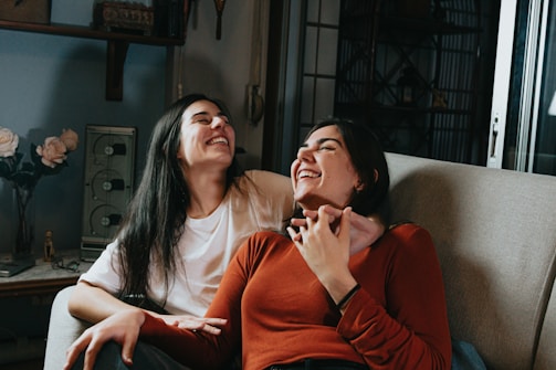 Two people sitting on a couch are sharing a joyful moment, laughing together. One is wearing a white t-shirt while the other is in a rust-colored sweater. The setting appears to be indoors with a cozy atmosphere, as decorative elements like flowers and a vintage radio are visible in the background.