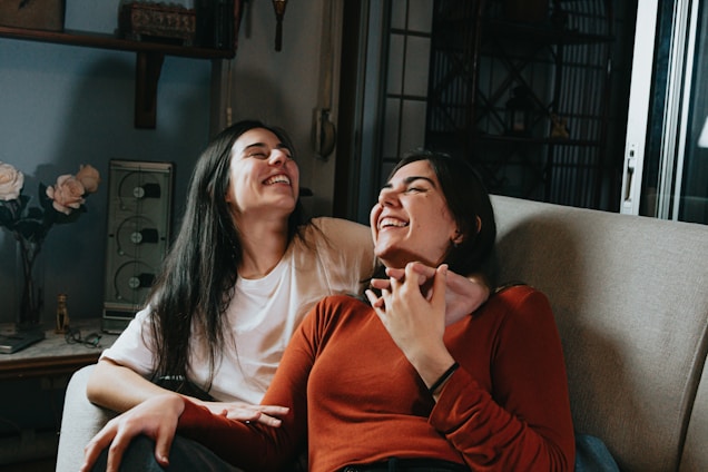 Two people sitting on a couch are sharing a joyful moment, laughing together. One is wearing a white t-shirt while the other is in a rust-colored sweater. The setting appears to be indoors with a cozy atmosphere, as decorative elements like flowers and a vintage radio are visible in the background.