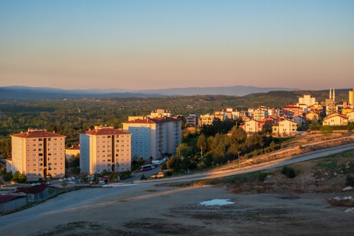A tranquil landscape with residential buildings and lush greenery bathed in warm evening light. The foreground features several mid-rise apartment buildings, while a sprawling town extends into the distance, surrounded by rolling hills.