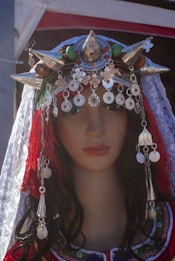 A mannequin head is adorned with a traditional and elaborate headpiece featuring metallic ornaments and conical shapes. The headpiece includes silver coins and intricate designs, with a veil that has lace detailing draped over the mannequin. The mannequin's hair is long and dark, styled in loose waves.