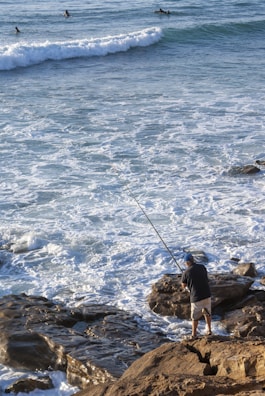 A person stands on rocky shore with a fishing rod, casting into the ocean. The sea is turbulent with waves crashing onto the rocks. In the background, several surfers are seen paddling and waiting for waves.