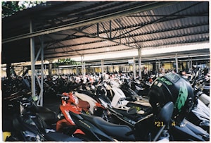 A crowded parking area filled with numerous motorcycles lined up under a large covered structure. The roof is supported by metal beams and poles. There is a variety of motorcycle models and colors, with a prominent helmet placed on one of the motorcycles.