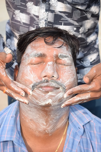 A man with closed eyes has shaving cream applied to his face by another person. The man wears a blue plaid shirt, while the other person is dressed in a patterned dark shirt and is carefully applying the cream.