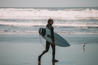A surfer in a wetsuit walks across the sandy beach while carrying a white surfboard under one arm. The ocean waves crash in the background, and a seagull stands nearby on the wet sand, reflecting the overcast sky.