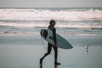 A surfer in a wetsuit walks across the sandy beach while carrying a white surfboard under one arm. The ocean waves crash in the background, and a seagull stands nearby on the wet sand, reflecting the overcast sky.