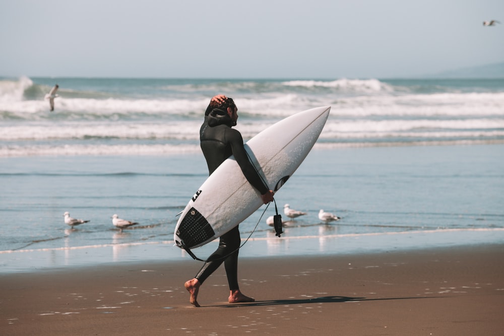 woman in black wetsuit holding white surfboard walking on beach during daytime