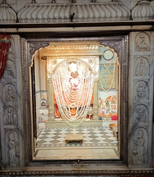 An ornately decorated deity inside a temple, adorned with garlands and intricate carvings on the surrounding stonework. The statue is centered within an elaborate altar, framed by detailed marble engravings. The floor in front has a checked pattern, and the scene is lit with a warm glow, highlighting the rich colors and textures.