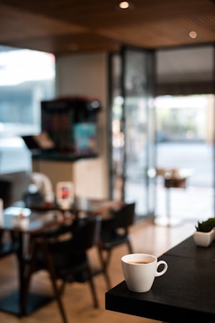 A cozy cafe with a modern interior featuring wooden paneling on the ceiling. A small white coffee cup, filled with coffee, is placed in focus on a black tabletop in the foreground. The background shows blurred furniture, including tables and chairs, along with a doorway leading outside.