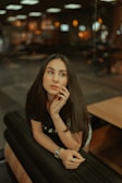 A thoughtful woman with long dark hair, leaning on a cushioned surface, appears to be lost in contemplation. Her hand rests near her face, and she's wearing a black t-shirt. The background is softly blurred with warm lighting, suggesting a cozy indoor space.