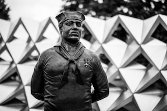 A monochrome sculpture of a sailor with a cap and a medal pinned to his chest. The background features a geometric pattern of interconnected triangular shapes.