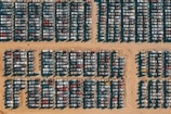 Rows of cars are tightly packed in a grid pattern on a sandy surface, likely a junkyard or storage lot. The harsh sunlight creates distinct shadows that highlight the arrangement of the vehicles.
