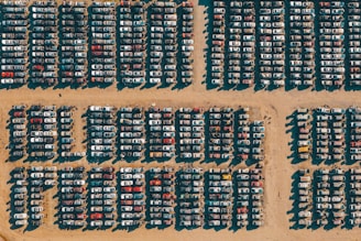 Rows of cars are tightly packed in a grid pattern on a sandy surface, likely a junkyard or storage lot. The harsh sunlight creates distinct shadows that highlight the arrangement of the vehicles.