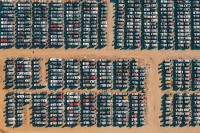 Rows of cars are tightly packed in a grid pattern on a sandy surface, likely a junkyard or storage lot. The harsh sunlight creates distinct shadows that highlight the arrangement of the vehicles.