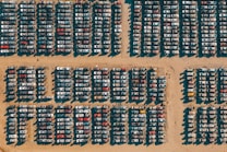 Rows of cars are tightly packed in a grid pattern on a sandy surface, likely a junkyard or storage lot. The harsh sunlight creates distinct shadows that highlight the arrangement of the vehicles.