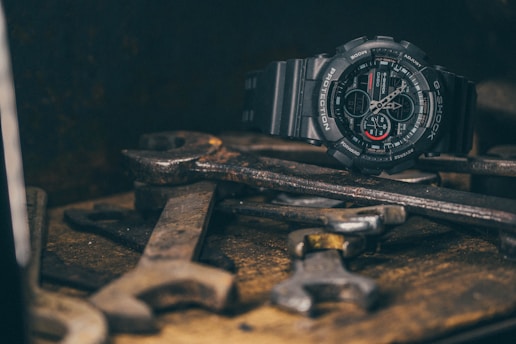 A rugged, black G-Shock watch is placed on top of a pile of old, rusty wrenches. The scene has a vintage and industrial feel, with the watch's dark and modern design contrasting against the worn tools.