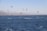 Numerous kiteboarders are gliding across a vast expanse of water, with colorful kites soaring in the sky above them. In the distance, there is a coastline with mountains and some industrial structures visible.