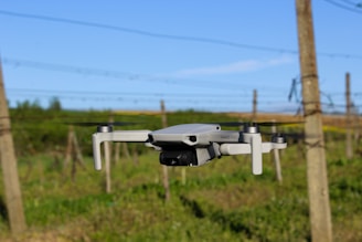 A drone hovers in mid-air against the backdrop of a rural landscape. The scene captures posts and wires in the background, likely indicating the edge of a field or vineyard. The sky is clear with an expansive blue hue.