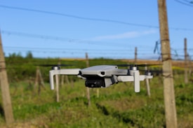 A drone hovers in mid-air against the backdrop of a rural landscape. The scene captures posts and wires in the background, likely indicating the edge of a field or vineyard. The sky is clear with an expansive blue hue.
