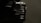 A close-up view of a black, hard-shell suitcase with a ribbed texture, partially in shadow. The suitcase appears to be sturdy, with metal rivets and reinforced edges. The lighting creates a dramatic effect, highlighting certain parts while leaving others in darkness.