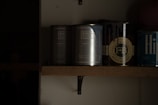A dimly lit shelf displaying several cans of paint. The labels indicate brands such as Porter's Paints and Farrow & Ball. The cans are organized in a row, with some text partially visible due to the lighting.