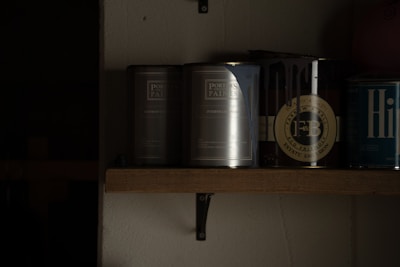 A dimly lit shelf displaying several cans of paint. The labels indicate brands such as Porter's Paints and Farrow & Ball. The cans are organized in a row, with some text partially visible due to the lighting.