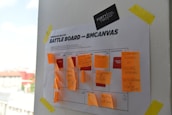 A business model canvas titled 'Battle Board' is taped to a white wall with yellow tape. The canvas is covered with orange sticky notes containing handwritten text. The wall card at the top says 'Forty Two.” Outside the window, blurred buildings are visible under a blue sky with white clouds.